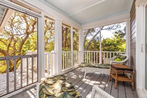 A covered porch with a white railing, a wicker chair with a green cushion, and a wicker table. - Spacious! 3 Carts-Captain's Watch Views-Steps to W Beach-4 day summer min stay (Bald Head Island)