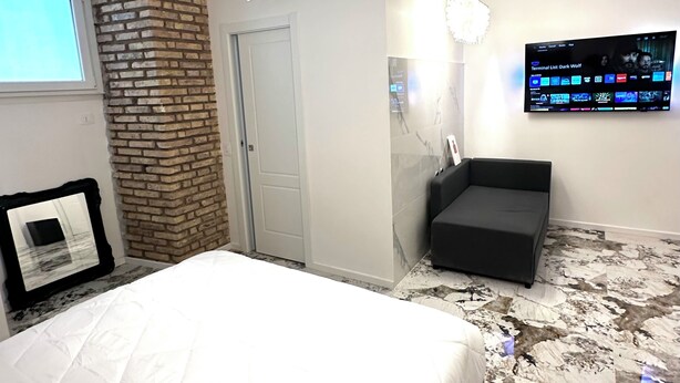 A modern bedroom with a large bed, a brick accent wall, a mounted TV, and a unique chandelier. - Callas Luxury Apartment (Roma)