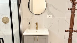 A bathroom with a glass shower enclosure, a white sink with gold fixtures, a round mirror, and a wooden towel rack. - Callas Luxury Apartment (Roma)