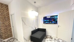 A modern room with a marble-patterned floor, a black sofa, a brick accent wall, and a mounted TV displaying a film. - Callas Luxury Apartment (Roma)