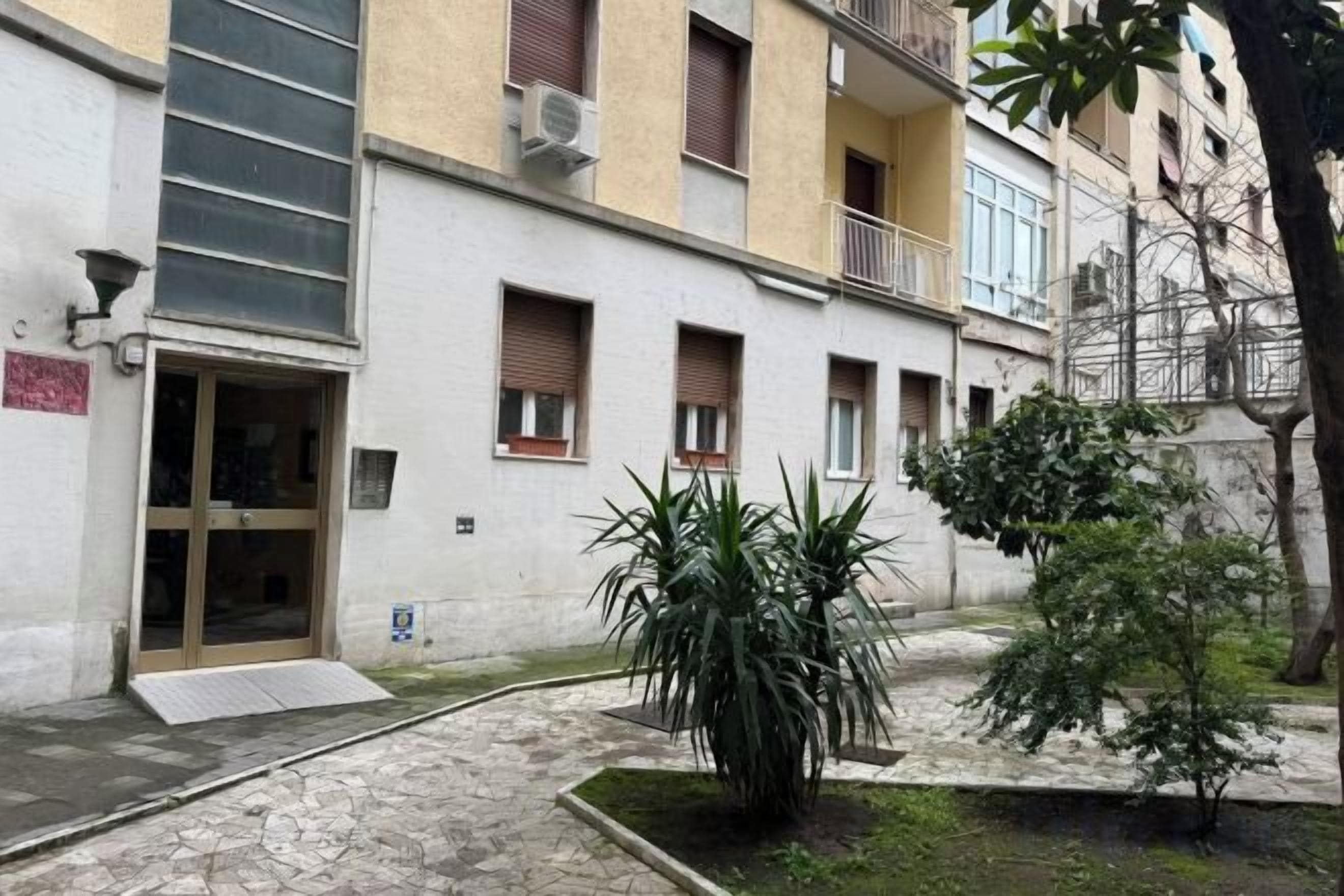 A building with a ground entrance, a small garden area with plants, and a paved walkway.