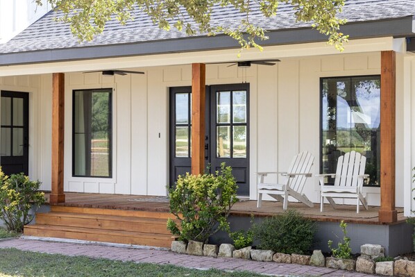 Una casa moderna con un porche cubierto, dos sillas Adirondack blancas y un pequeño jardín.