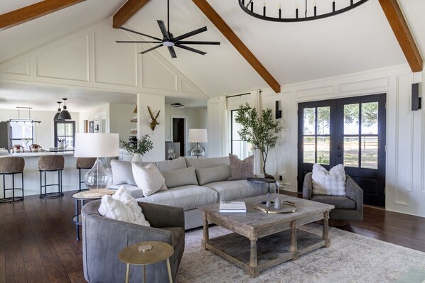 A spacious living room with a large gray sofa, a coffee table, and a wooden bench. There is a kitchen in the background with bar stools. - Windy Hill Retreat | Modern Farmhouse with Views, Cowboy Pool &amp; Firepit (La Grange)