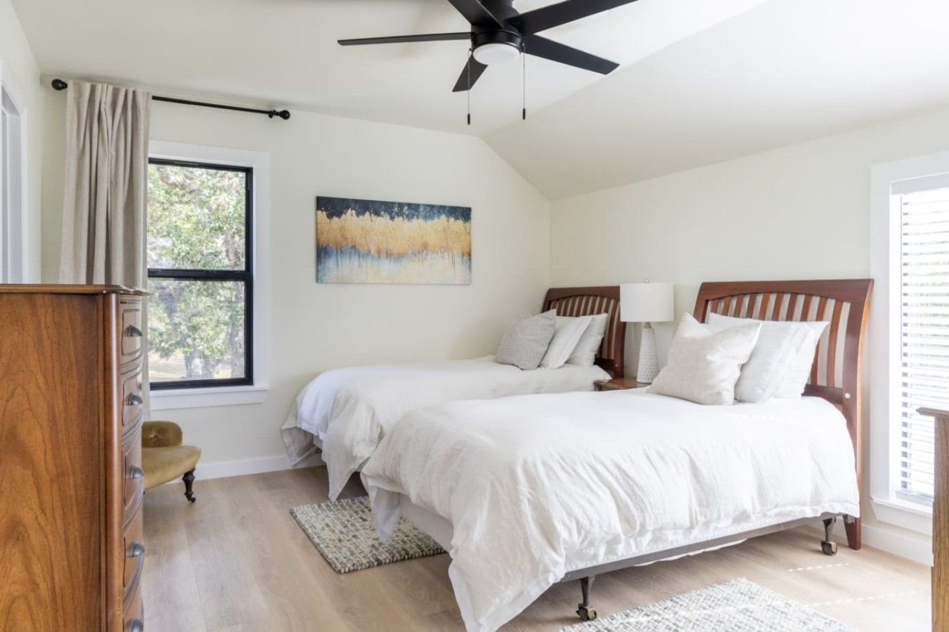Quarto com duas camas, um ventilador de teto, janela com cortinas, um quadro na parede e uma cômoda de madeira.