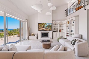 A spacious living room with a large sofa, a coffee table, and a fireplace. There is a television mounted above the fireplace and a bookshelf to the right. The room has large windows with a view of the outdoors.