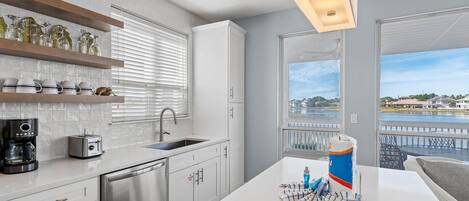 A modern kitchen with white cabinetry, a stainless steel dishwasher, a coffee maker, and a toaster. There is a window with blinds, a view of a body of water, and a balcony with a railing.