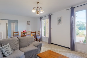 Een woonkamer met een bank, een houten salontafel, een eethoek met stoelen en een tafel, en een groot raam met gordijnen.
