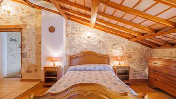 A bedroom with a wooden bed, stone walls, a sloped ceiling with wooden beams, and a dresser.