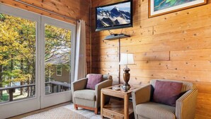 Una habitación acogedora con paredes de madera, un ventanal con vistas a árboles, dos sillones a rayas, una mesita con lámpara y un televisor montado.