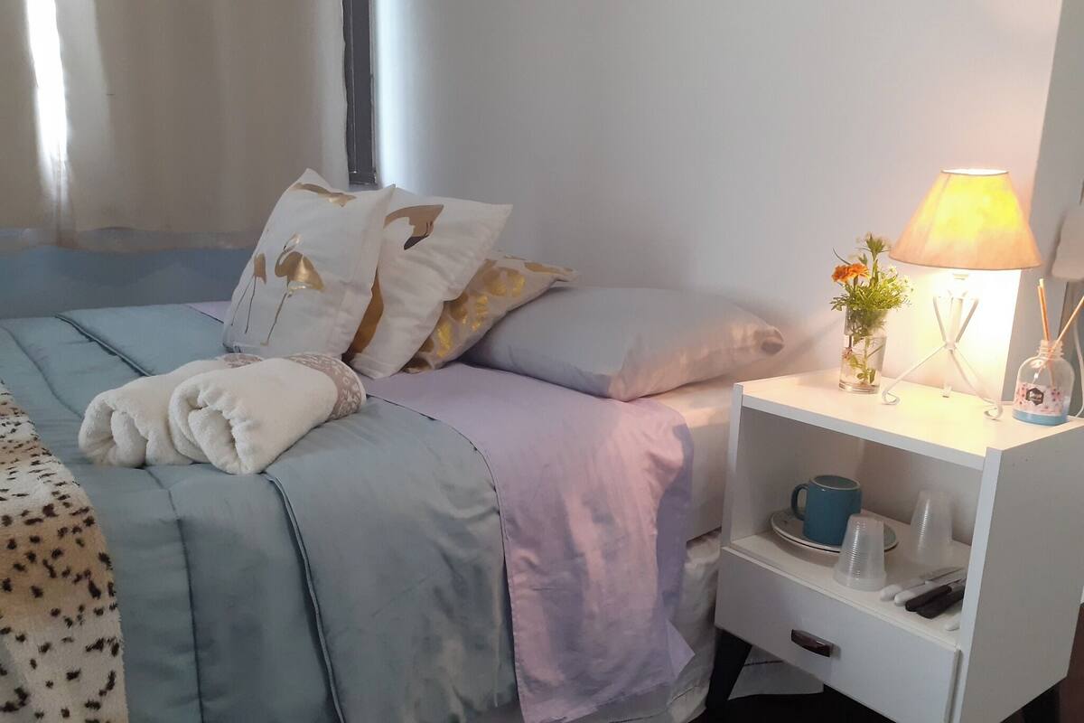 Un lit bien fait, avec une couette bleue et blanche, un oreiller blanc orné d’un motif doré et une serviette blanche. Sur la table de chevet, on trouve une lampe, un vase avec des fleurs et une petite étagère sur laquelle sont posés une tasse et une bouteille.