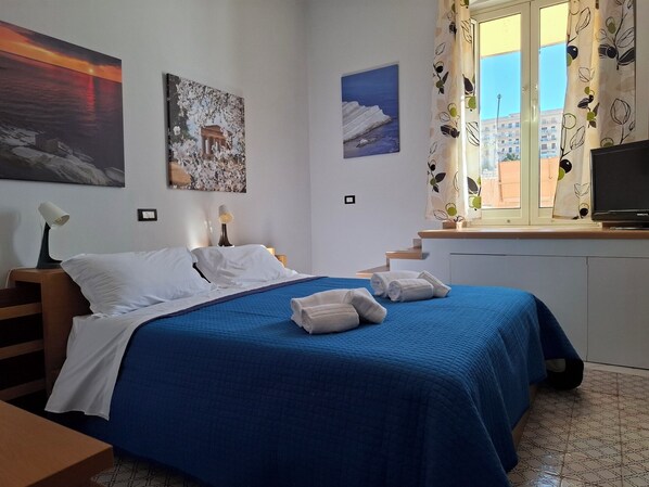 Une chambre à coucher avec un lit, deux lampes fixées au mur, une télévision et une fenêtre donnant sur des immeubles.