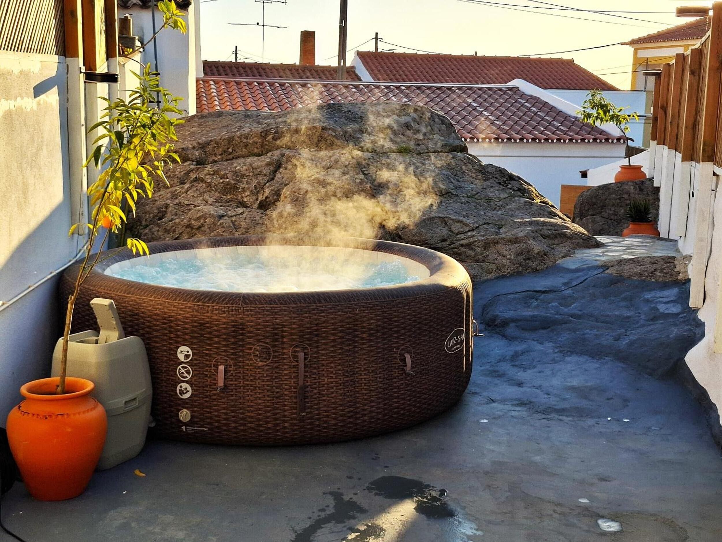 Un jacuzzi en un patio con una roca grande, una planta en maceta y un pequeño jarrón naranja.