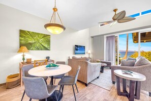 A modern living room with a round dining table, grey sofas, a ceiling fan, and a large window with a view of the ocean.