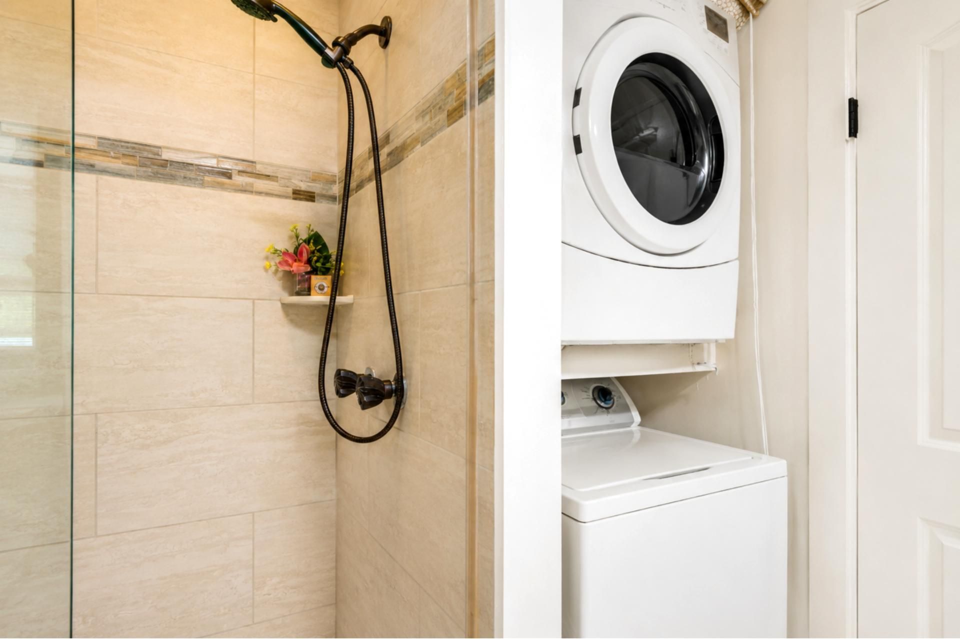 A bathroom with a shower, a washing machine, and a small shelf with flowers.