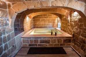 A stone-walled hot tub with a rectangular shape, two bottles of champagne, and a black mat at the entrance.