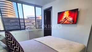 A modern bedroom with a large window, a bed with a patterned pillow, and a wall-mounted TV displaying a fiery image.