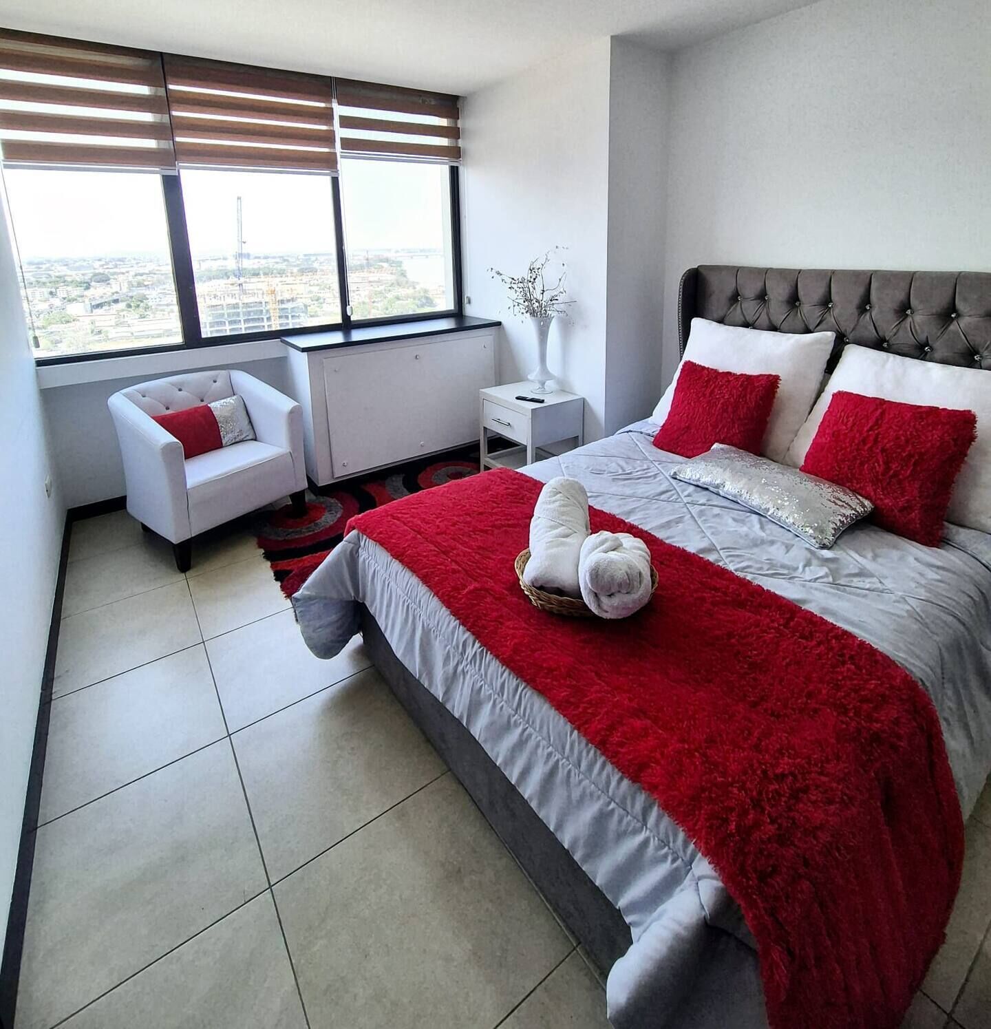 Une chambre moderne avec un grand lit, un fauteuil blanc, une table de chevet et une fenêtre avec des stores.
