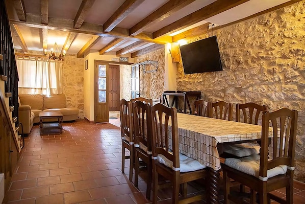 A dining area with a long wooden table and chairs, a television mounted on a stone wall, and a sign indicating the exit.