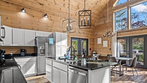 A modern kitchen with white cabinets, a stainless steel refrigerator, and a granite countertop. There is a dining area with a table and chairs, and large windows providing natural light. - New! Luxe Pool Cabin w/Hot Tub*Theater*Game Room (Gatlinburg)
