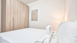 Un lit bien fait, avec du linge de lit blanc et une serviette enroulée, une armoire en bois et un petit tableau encadré accroché au mur.