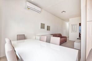 A modern, minimalist living room with a white dining table, a sofa, and a small kitchen area. - Mercury Residence No.15 / Penthouse / Terrace (Sliema)