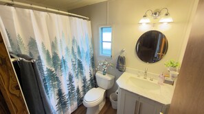 A bathroom with a shower curtain featuring a forest print, a toilet, a sink with a mirror above it, and a window.