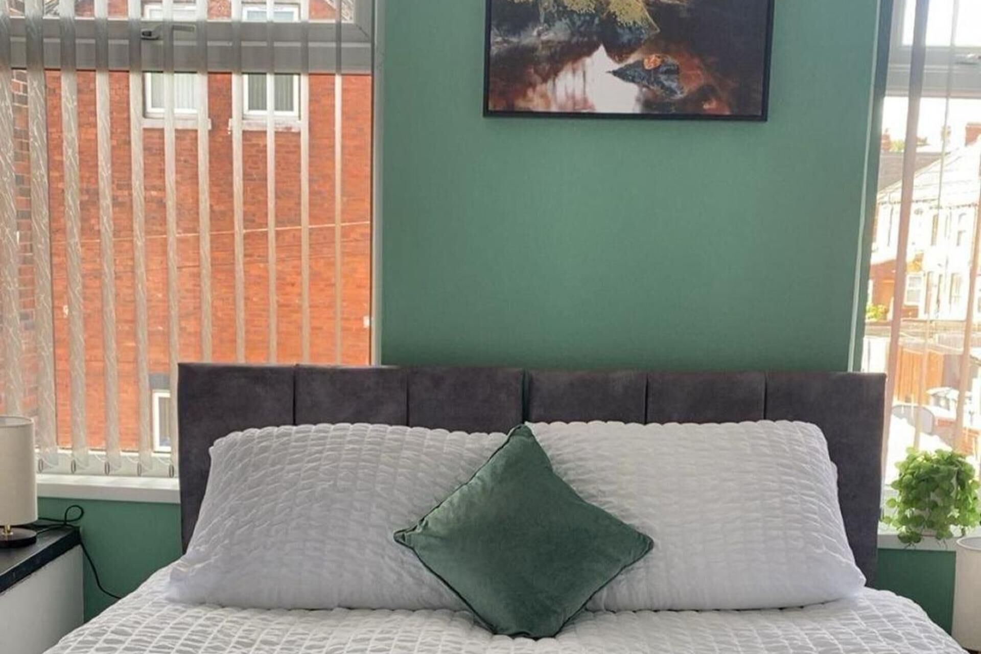 Un lit recouvert d’une couette blanche et orné d’un oreiller vert, un tableau accroché au mur et, par la fenêtre, une vue sur un bâtiment en briques.