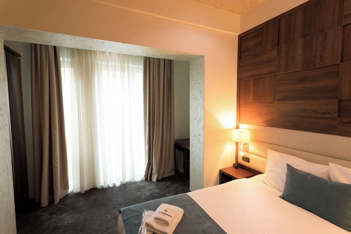 Una camera d'albergo con un letto, un comodino con una lampada, una finestra con tende e una testiera in legno.