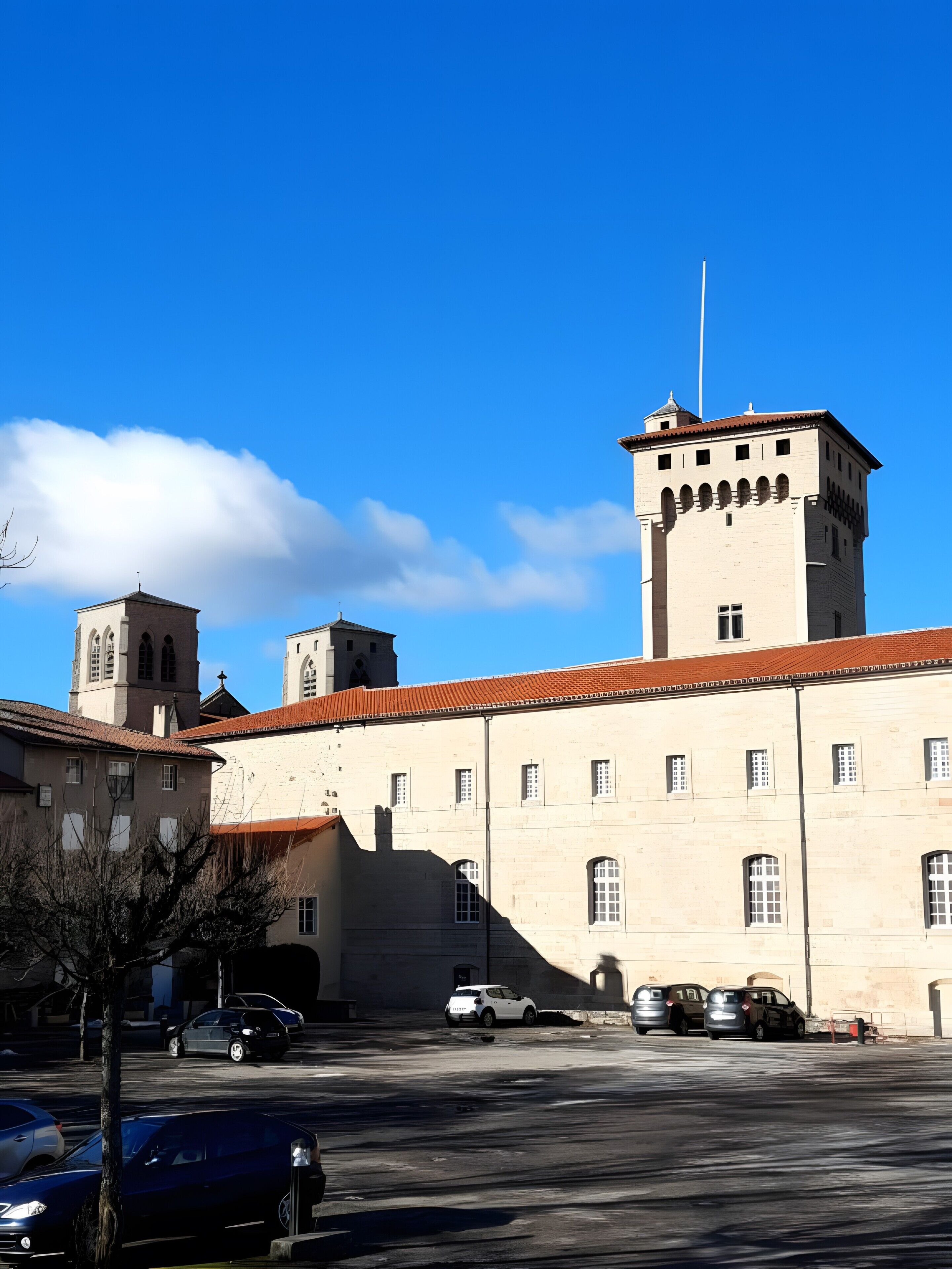 A large building with a prominent tower, multiple windows, and a parking area with cars.