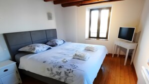 Une chambre à coucher comprenant un lit, un téléviseur, un plancher en bois, une fenêtre et un plafond avec des poutres apparentes.