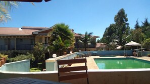 A pool area with a wooden bench, palm trees, and a building with a tiled roof. - Eden Springs Hotel (Kapsabet)