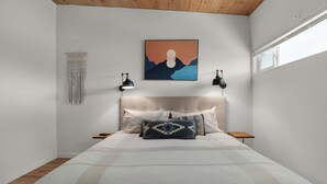A bedroom with a large bed, two bedside tables, a wall-mounted lamp, and a framed artwork above the bed.