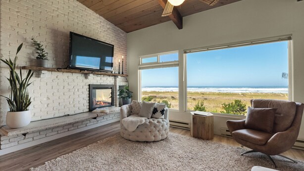A living room with a fireplace, a TV mounted on the wall, and a large window overlooking the beach.