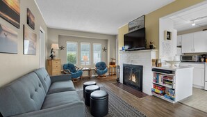 A modern living room with a grey sofa, blue chairs, a fireplace, and a kitchen area with white cabinets and a TV. - The Tides by the Sea - Entire rental unit in Seaside, Oregon, United States (Seaside)