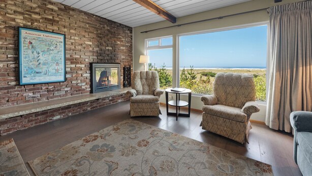 A room with a brick wall, a fireplace, two armchairs, a small table, and a view of the beach.