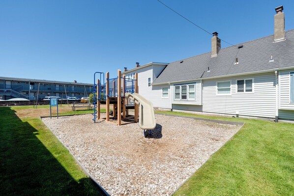 Un parque infantil con un tobogán y estructuras para escalar, rodeado de grava y césped, con un edificio al fondo.