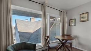 Quarto com vista para edifícios vizinhos, uma poltrona verde, uma mesa de jantar de madeira com duas cadeiras e dois quadros na parede.