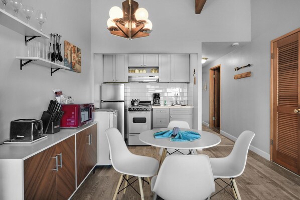A modern kitchen with white cabinets, a dining area with white chairs, and a wooden door. - The Tides by the Sea - Entire rental unit in Seaside, Oregon, United States (Seaside)