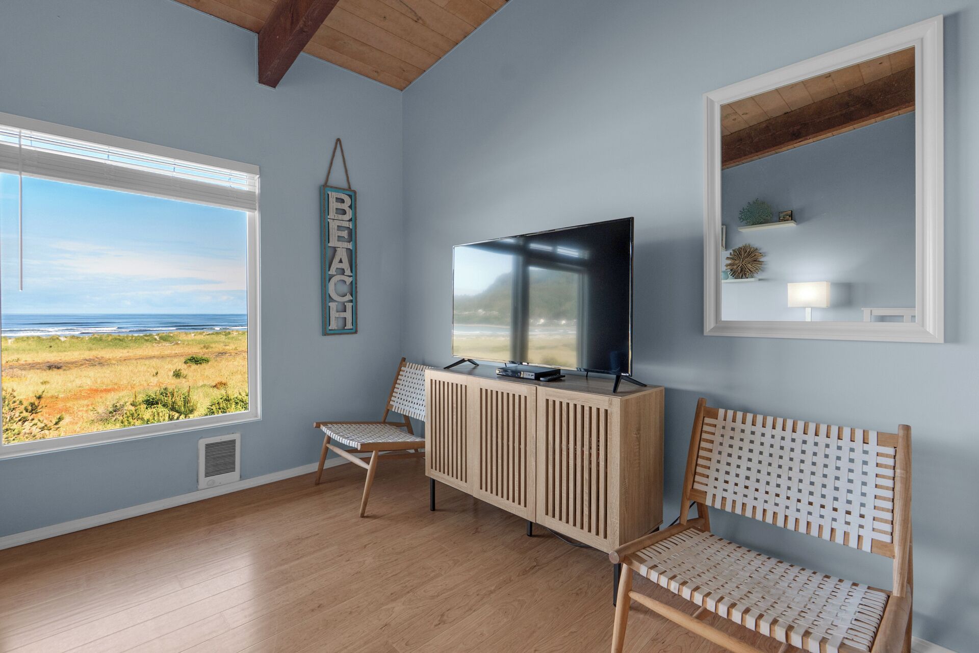 Ein Zimmer mit großem Fenster und Blick auf den Strand, einem Fernseher auf einer Holzkommode und einem kleinen Regal mit Dekorationsgegenständen.