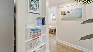 A hallway with a white wall, a shelf with books and decorative items, and a framed picture of a beach scene.