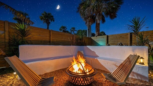 A night scene with a fire pit, two lounge chairs, and a wooden fence.