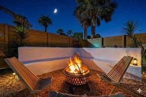 Une scène nocturne avec un foyer, deux fauteuils de jardin et une clôture en bois.