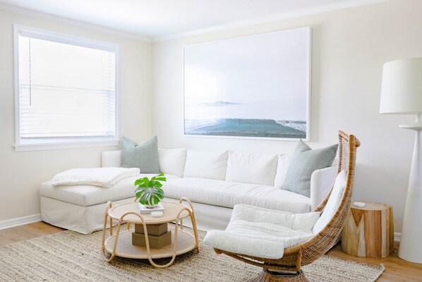 Ein Wohnzimmer mit einem weißen Sofa, einem Korbstuhl, einem runden Tisch mit einer Pflanze und einem großen Fenster mit Meerblick.