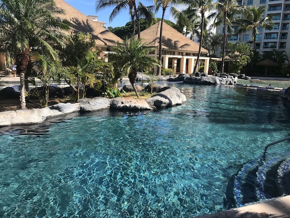 Uma piscina cristalina com pedras, palmeiras e um edifício com telhado de palha ao fundo.