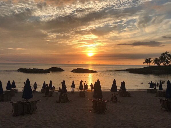 Ein Strand mit Sonnenschirmen und Tischen, ein Sonnenuntergang und ruhige See.
