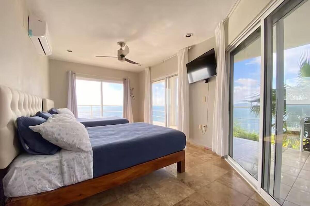 Un dormitorio con una cama grande, un ventilador de techo, un televisor montado en la pared y vistas al océano a través de amplias puertas correderas.