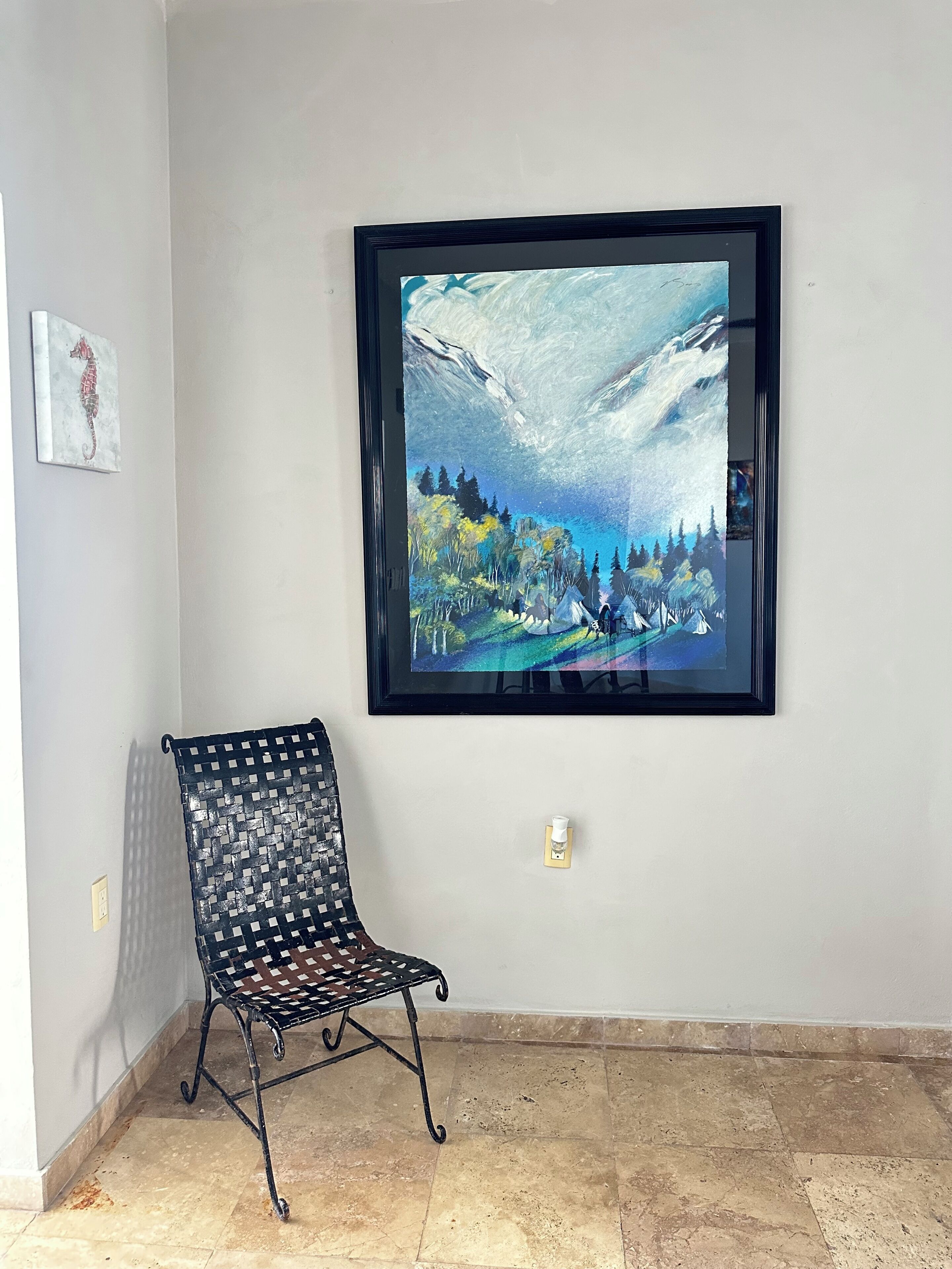 A framed artwork of a landscape with mountains and trees, a black metal chair, and a tiled floor.