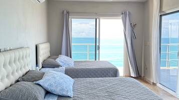 A bedroom with two beds, a ceiling fan, an air conditioning unit, and a view of the ocean through a sliding glass door.