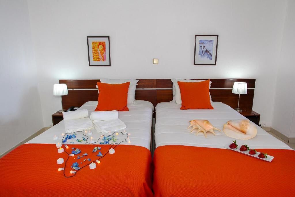 A hotel room with two beds, each with an orange bedspread, white linens, and matching orange pillows. There are framed pictures on the wall, a nightstand with a lamp, and a tray with strawberries and a banana on the beds.