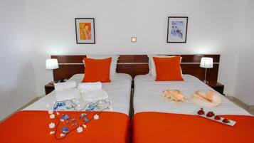A hotel room with two beds, each with an orange bedspread, white linens, and matching orange pillows. There are framed pictures on the wall, a nightstand with a lamp, and a tray with strawberries and a banana on the beds.